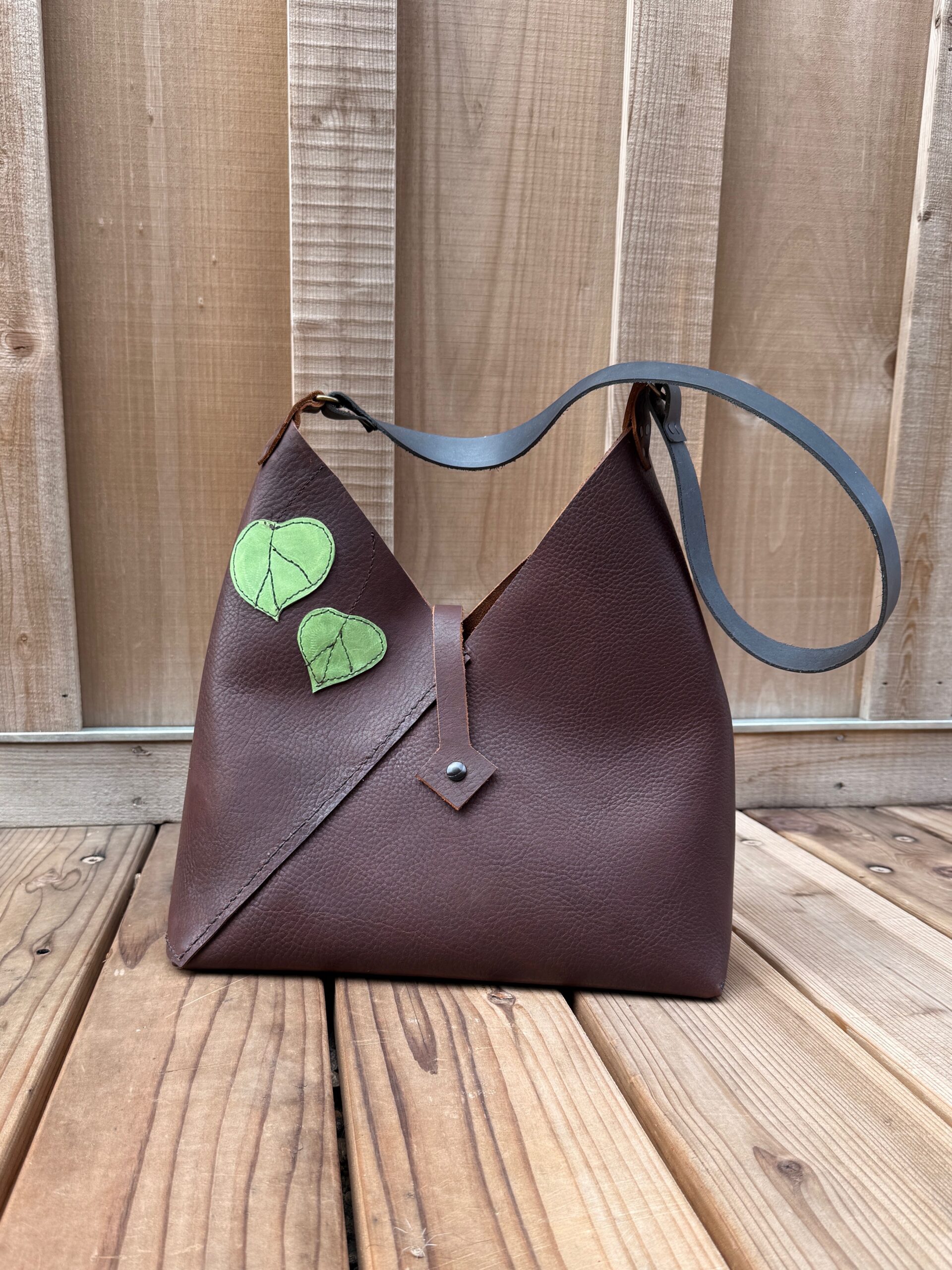Handmade dark brown leather shoulder bag with green stitched leaf appliqués, black leather strap, and asymmetrical design.