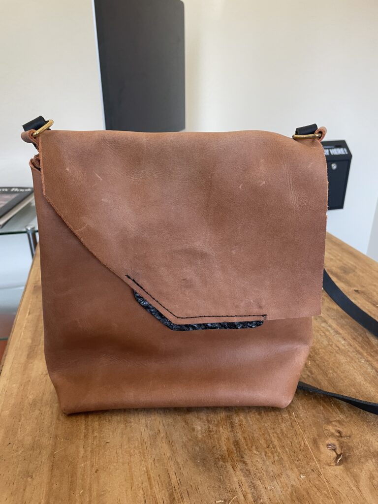 Handmade brown leather messenger bag with asymmetrical flap, black accent detail, brass hardware, and shoulder strap.