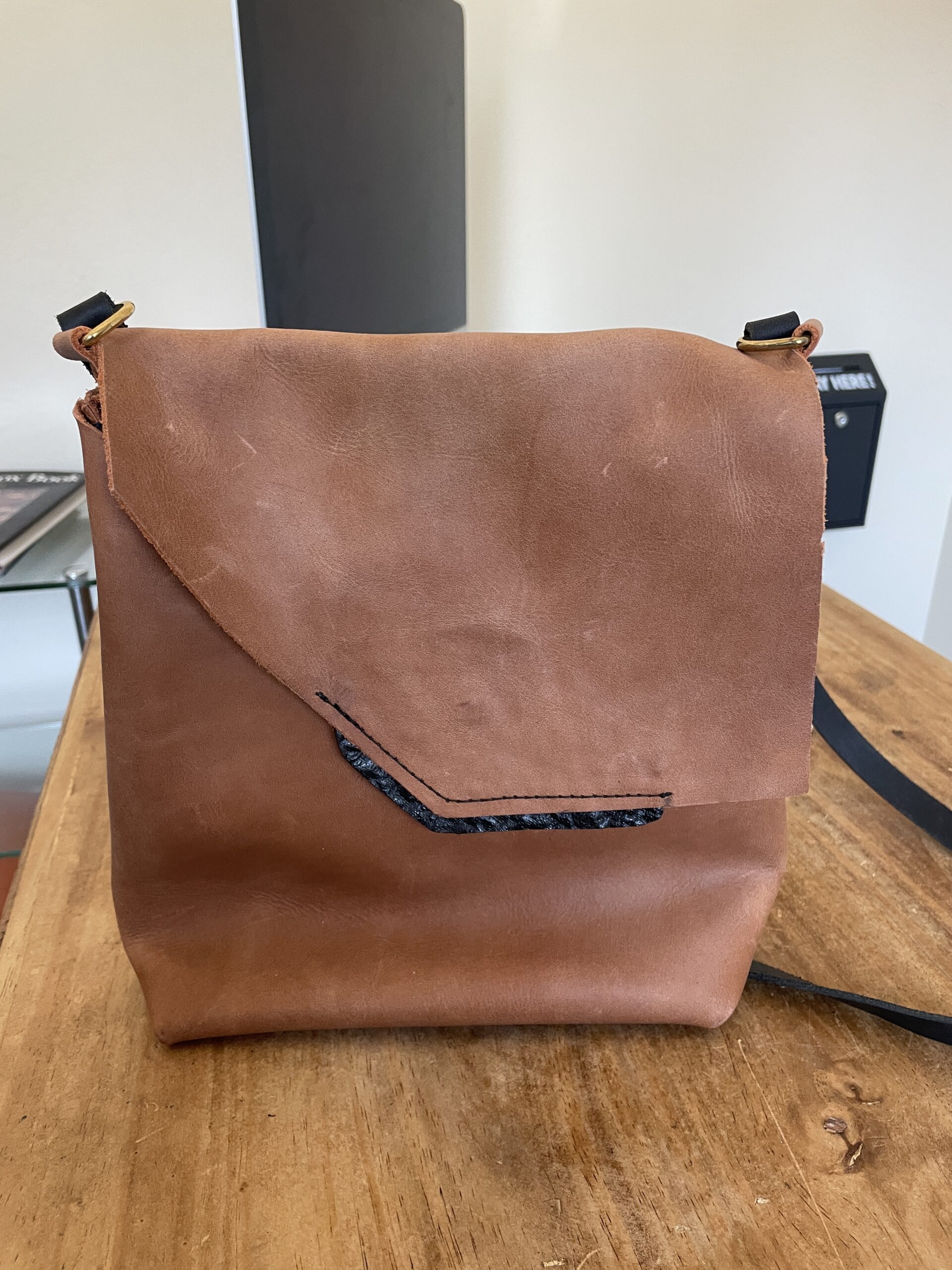 Handmade brown leather messenger bag with asymmetrical flap, black accent detail, brass hardware, and shoulder strap.