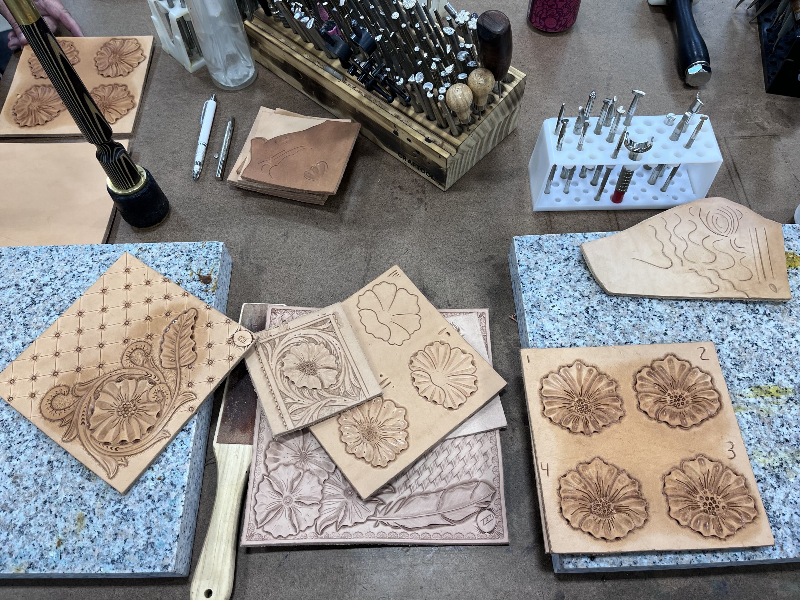 Hand-tooled leather samples featuring floral patterns, feathers, and decorative designs surrounded by leatherworking tools at Rocky Mountain Folk School in Grand Lake, Colorado.